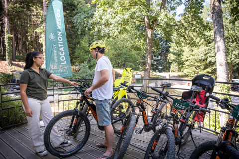 Dos personas en el área de alquiler de bicicletas en Huttopia Wattwiller rodeadas de bicis y naturaleza verde.