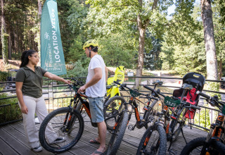 Due persone nella zona noleggio bici di Huttopia Wattwiller, con mountain bike e scenario boschivo.