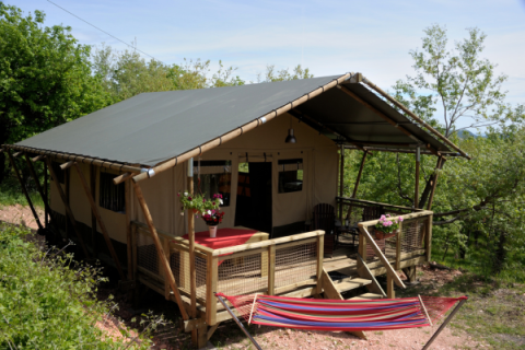 Tienda safari Wood Lodge en Camping Casa Tartufo en Italia, con terraza de madera y hamaca entre árboles.
