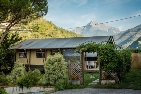 Tienda safari Wood Lodge en Camping Delle Rose, Italia, rodeada de vegetación y montañas al fondo.
