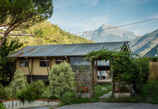 Tienda safari Wood Lodge en Camping Delle Rose, Italia, rodeada de vegetación y montañas al fondo.