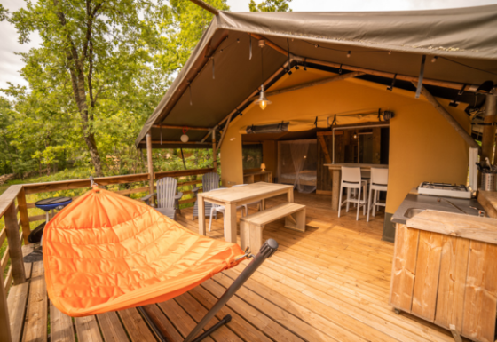 Tenda Safari Lodge con terrazza in legno, amaca arancione, area pranzo e cucina all’aperto nella natura.