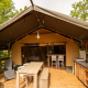 Safari Lodge safari telt med udendørs spisebord, træstole og køkken midt i frodige grønne omgivelser.