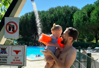 Een vader geeft zijn kindje met zwembandjes een douche naast het buitenzwembad onder een blauwe hemel in Frankrijk.