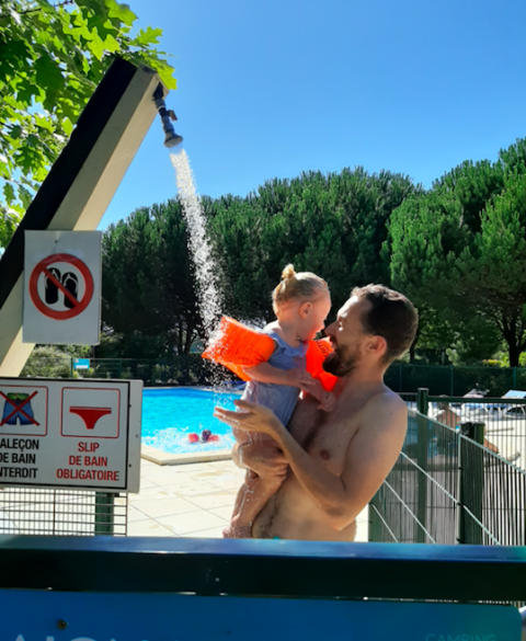 Un padre ducha a su hijo pequeño con manguitos junto a una piscina al aire libre bajo un cielo azul en Francia.