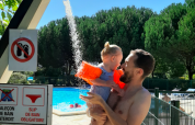 A father showers his small child with floaties near an outdoor pool under a bright blue sky in France.