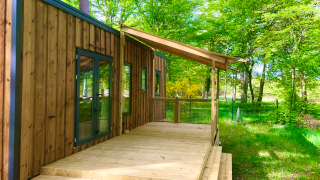 Cabaña Prestique de madera con porche cubierto rodeada de abundante vegetación y bosque verde en verano.