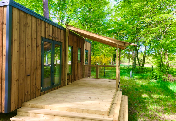 Cabaña Prestique de madera con porche cubierto rodeada de abundante vegetación y bosque verde en verano.