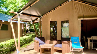 Safari tent with wooden deck, outdoor lounge chairs, and lush green surroundings in bright sunlight.