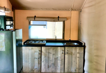 Zona cucina all'interno di una tenda safari con armadietti in legno, fornello a gas, lavello e frigorifero.