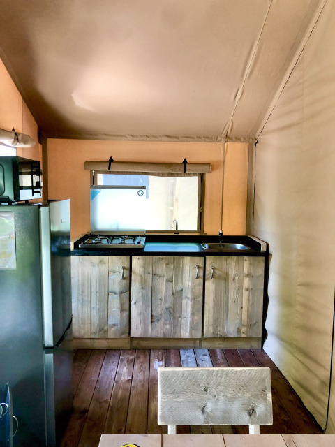 Coin cuisine dans une tente safari avec armoires en bois, gazinière, évier, réfrigérateur et sol en bois.