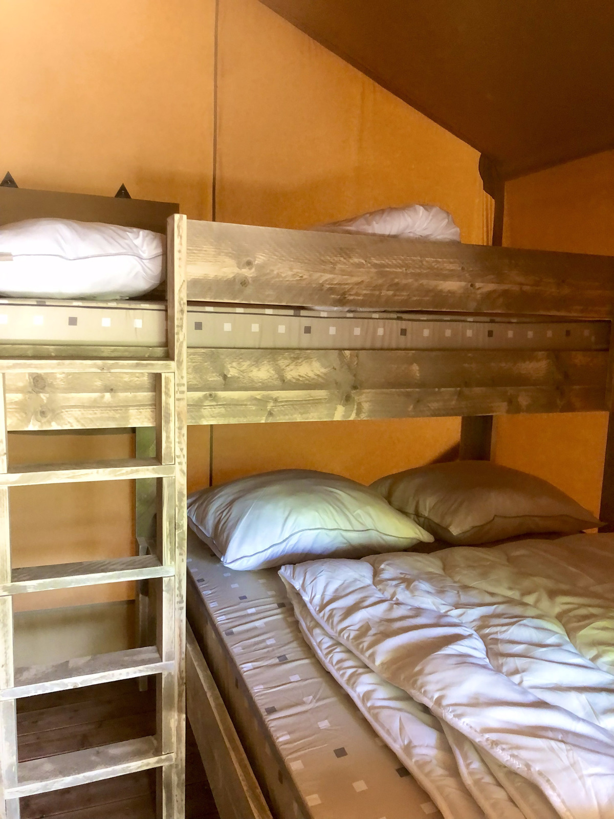 Houten stapelbed met kussens en dekbedden in een safaritent met warme, gele wanden.