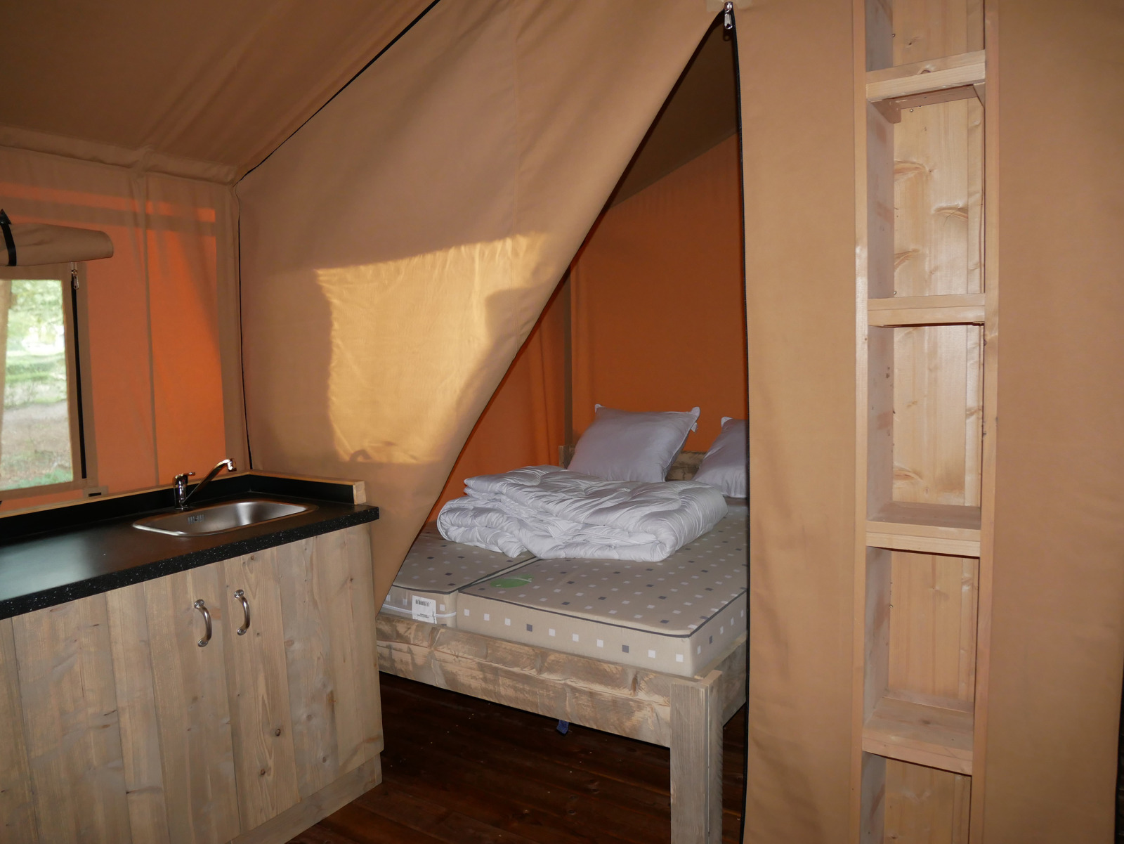 Binnenaanzicht van een safaritent met bed, houten planken en compacte kitchenette met spoelbak.