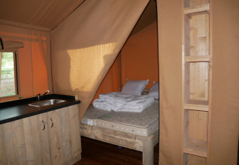 Vista interior de una tienda safari con cama, estanterías de madera y pequeña cocina con fregadero.