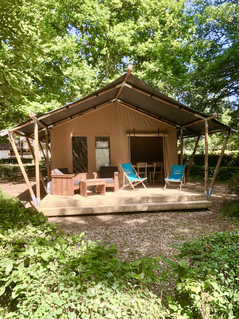 Tienda safari con terraza de madera, sillones y tumbonas, rodeada de vegetación y árboles frondosos.
