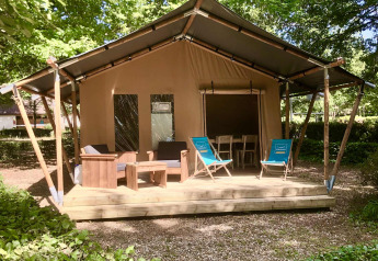 Tienda safari con terraza de madera, sillones y tumbonas, rodeada de vegetación y árboles frondosos.