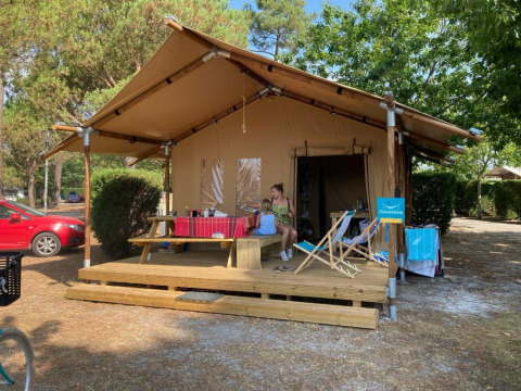 Tienda safari llamada Slow Lodge + baño, con dos personas sentadas en el porche, rodeadas de árboles.