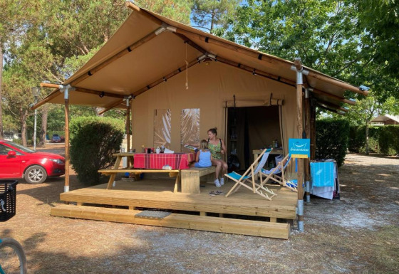 Tienda safari llamada Slow Lodge + baño, con dos personas sentadas en el porche, rodeadas de árboles.