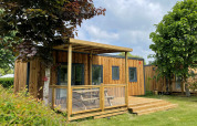 Prestige Cottage lodge in hout met terras, grasveld en omringd door bomen onder een bewolkte hemel.