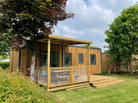 Lodge Prestige Cottage en bois avec terrasse, pelouse verte et arbres sous un ciel partiellement nuageux.