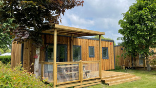 Lodge Prestige Cottage en bois avec terrasse, pelouse verte et arbres sous un ciel partiellement nuageux.