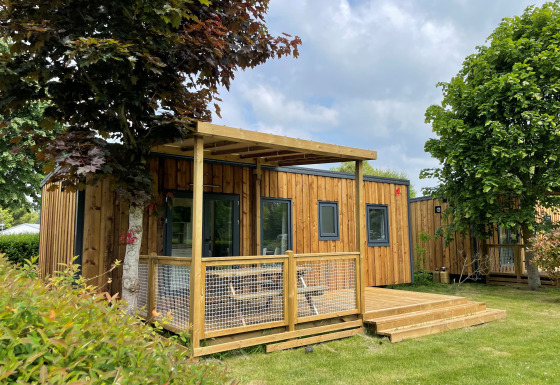 Lodge Prestige Cottage en bois avec terrasse, pelouse verte et arbres sous un ciel partiellement nuageux.
