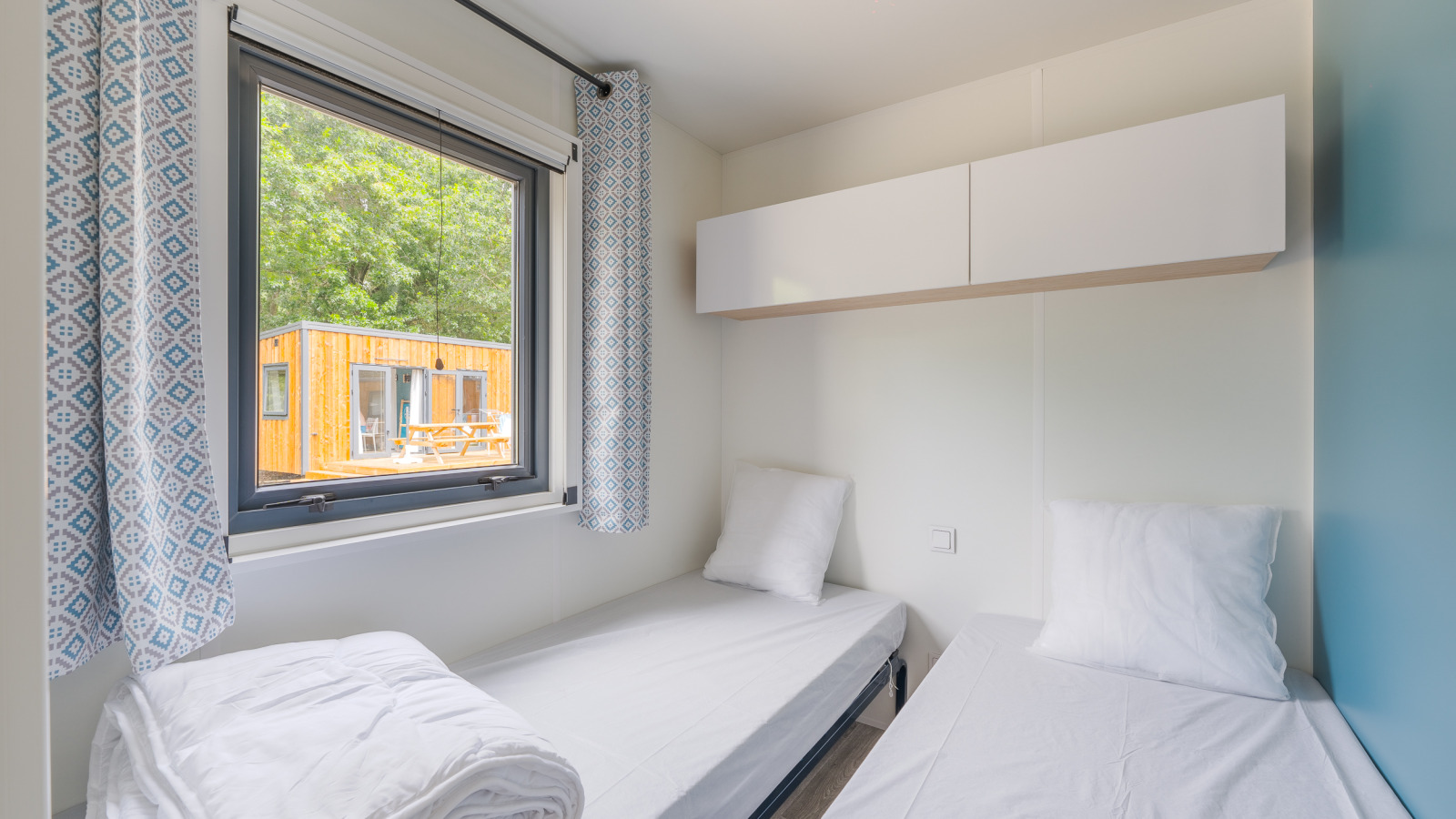 Twee eenpersoonsbedden in een lichte lodgekamer met raam, gordijnen en uitzicht op een houten huis buiten.