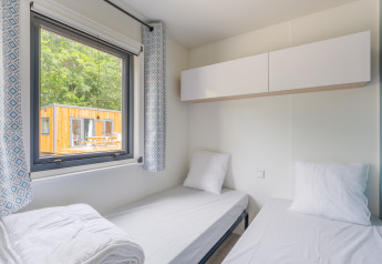 Twee aparte bedden in een helder lodge-kamertje met raam, gordijnen en zicht op een houten hut buiten.