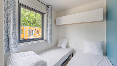 Twee eenpersoonsbedden in een lichte lodgekamer met raam, gordijnen en uitzicht op een houten huis buiten.