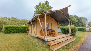 Tenda safari Slow Lodge con veranda in legno e mobili da esterno, circondata da siepi e alberi.
