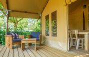 Outdoor seating area with wooden furniture on the porch of a safari tent surrounded by greenery.