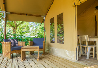 Gemütlicher Sitzbereich und Esstisch aus Holz auf einer Terrasse vor einem Safari-Zelt im Grünen.