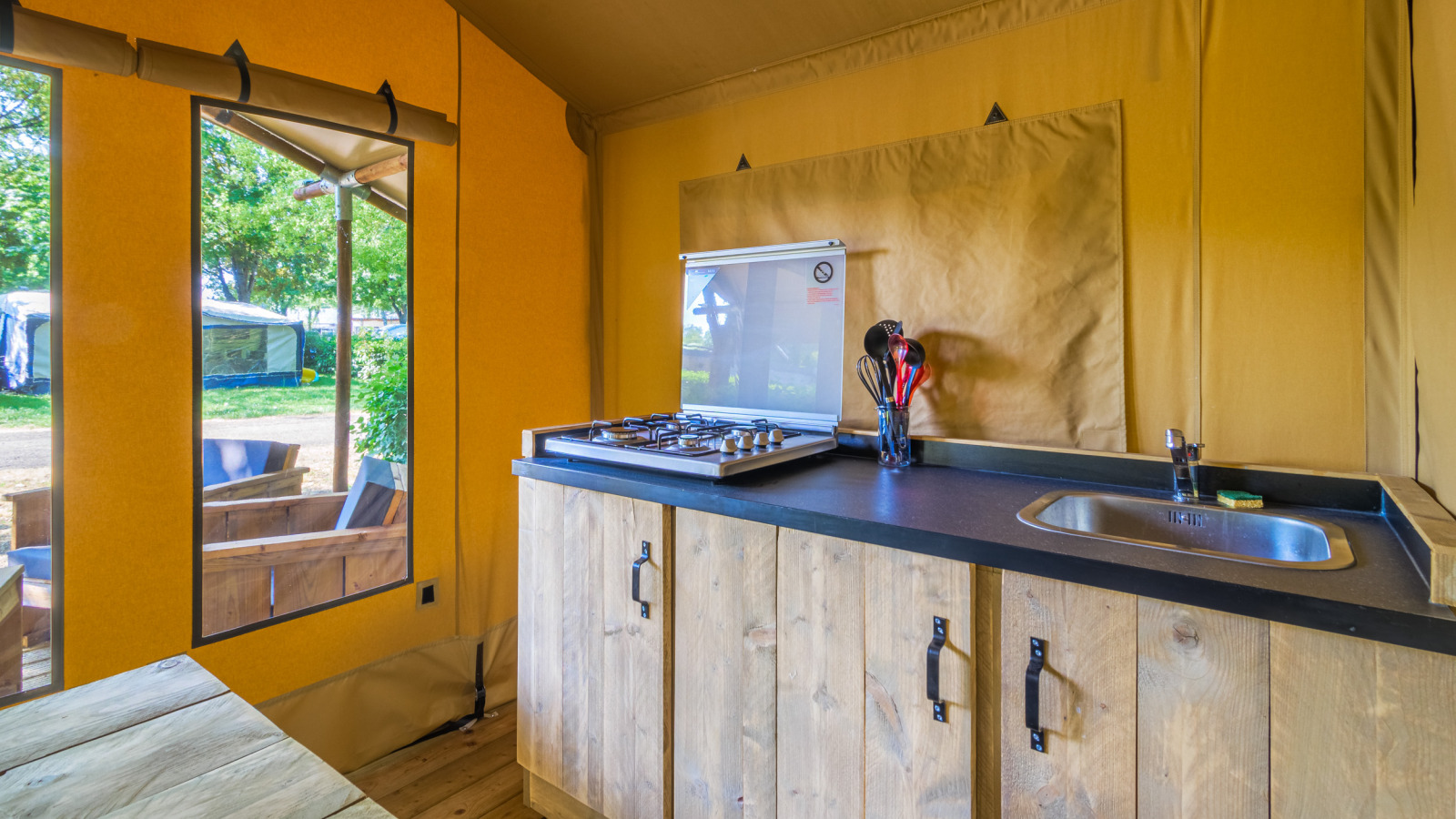 Interieur van een safaritent met houten keukenblad, gasfornuis, spoelbak en zicht op buitenzitje.