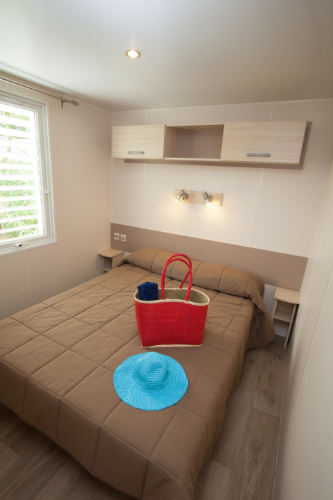 Slaapkamer in Cottage Soleo op Camping Seasonova Haliotis in Frankrijk met tweepersoonsbed, mand en hoed.
