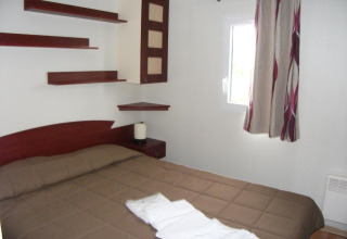 Schlafzimmer mit Doppelbett, Regalen, Fenster und Vorhängen in der Cottage Zen bei Camping Seasonova Haliotis.