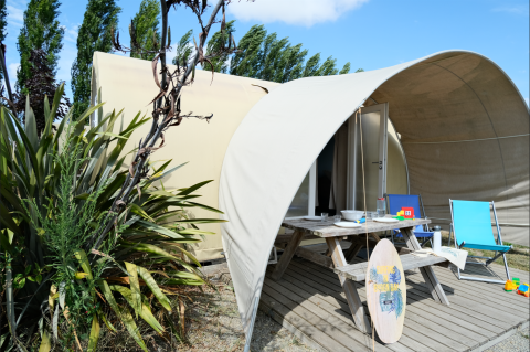 Tente safari au Slow Life Stay avec table de pique-nique, planche de surf et chaises, entourée de verdure.
