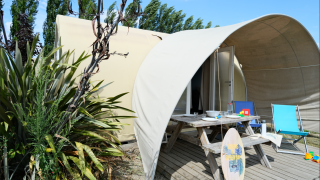 Safari-Zelt bei Slow Life Stay mit Picknicktisch, Surfbrett und Liegestühlen, umgeben von Pflanzen.