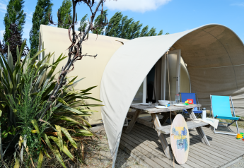 Safari-Zelt bei Slow Life Stay mit Picknicktisch, Surfbrett und Liegestühlen, umgeben von Pflanzen.