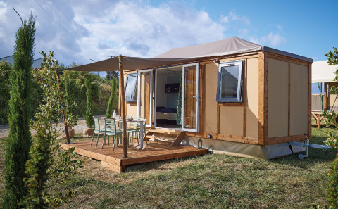 Vista exterior de la cabaña Corfou con terraza de madera, mesa, sillas y vegetación alrededor.
