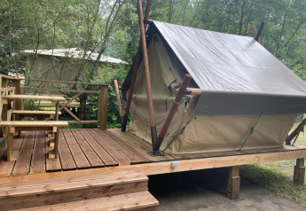 Bivouac teepee tent on elevated wooden deck with bench, surrounded by trees in a forest setting.