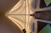 Vista interior de la tienda tipi Bivouac con dos camas, almohadas, mantas y una lámpara sobre piso de madera.