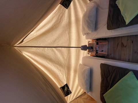 Vista interna del teepee Bivouac con due letti, cuscini, coperte e una lampada su pavimento in legno.