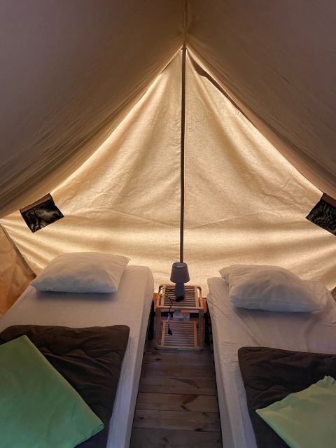 Vista interna del teepee Bivouac con due letti, cuscini, coperte e una lampada su pavimento in legno.