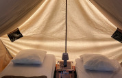Vista interna del teepee Bivouac con due letti, cuscini, coperte e una lampada su pavimento in legno.