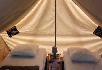 Vista interna del teepee Bivouac con due letti, cuscini, coperte e una lampada su pavimento in legno.