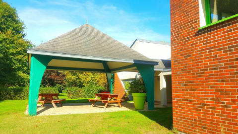 Outdoor covered picnic area with wooden benches, green pillars and brick building at Camping Seasonova Le Martinet.