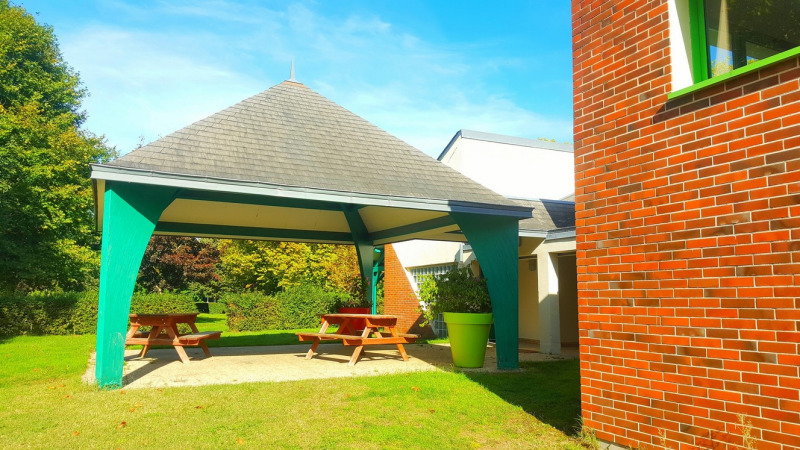 Overdekte picknickplaats met houten banken, groene pilaren en bakstenen gebouw bij Camping Seasonova Le Martinet.