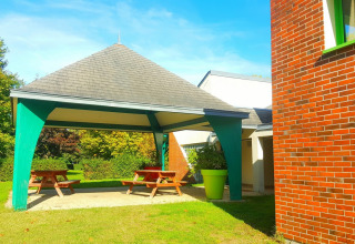 Outdoor covered picnic area with wooden benches, green pillars and brick building at Camping Seasonova Le Martinet.