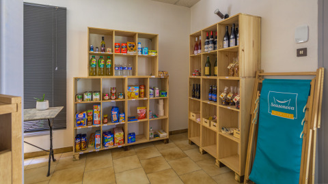 Indoor shop area with wooden shelves filled with groceries, wine bottles, snacks, and a blue Seasonova chair.