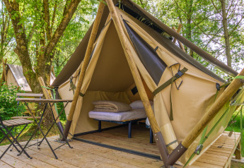A cozy glamping tent with a bed and small table set on a wooden deck surrounded by trees and greenery.
